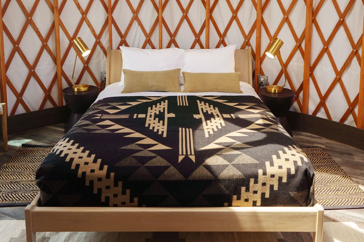 A bed in a yurt with a patterned blanket, two bedside lamps, and a wooden headboard.