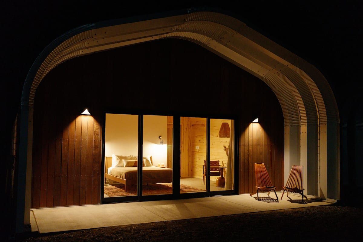 A modern hotel room with a large sliding glass door, a bed, a desk, and two chairs.