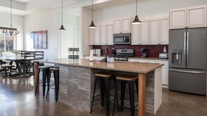 A modern kitchen with white cabinets, a large island with stools, a stainless steel refrigerator, and pendant lighting.