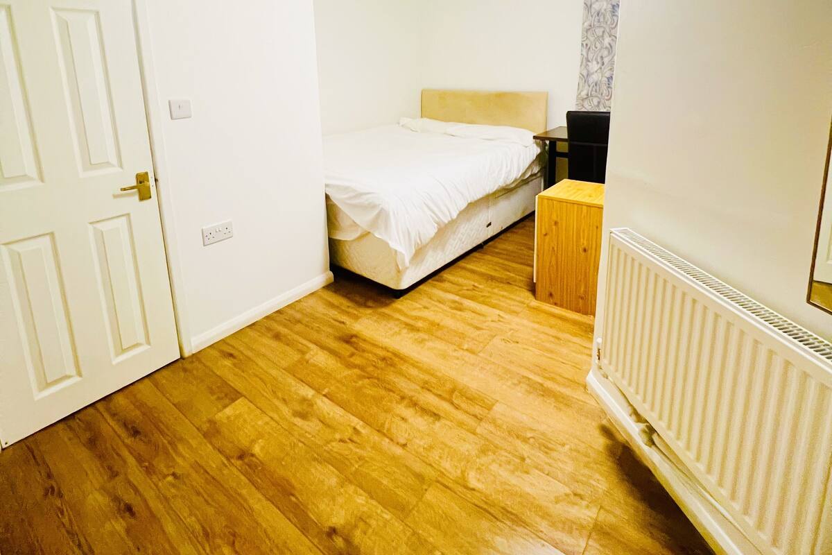 A bedroom with a wooden floor, a bed with a white headboard, a nightstand, and a radiator.