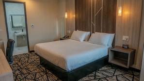 A hotel room with a large bed, bedside tables, a desk, a chair, a mirror, a vanity with a sink, and a patterned carpet. - Comfeeni Inn and Suites (South Houston)