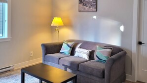 Un salon avec un canapé gris, une table basse en bois, une lampe, un tableau accroché au mur et une porte.