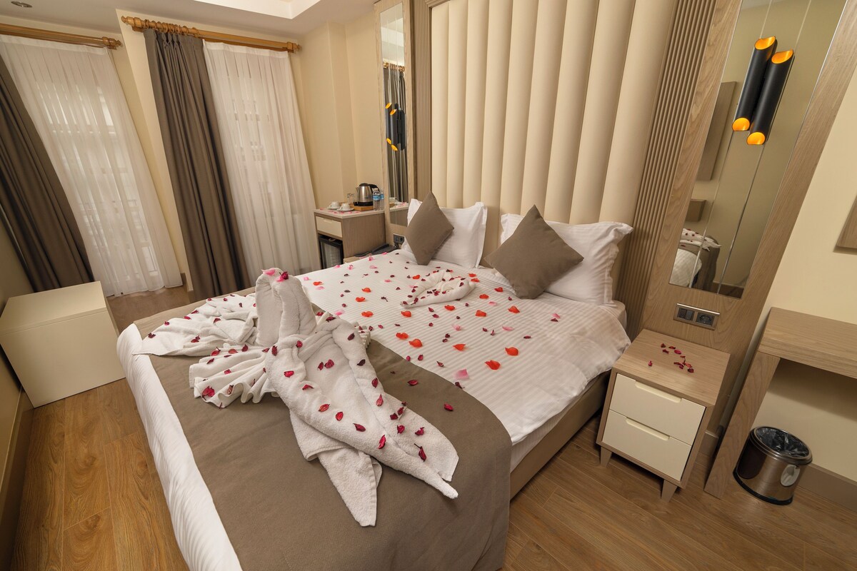 A neatly made bed with rose petals and a robe, a bedside table with a lamp, and a mirror with a wooden frame.