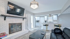 A modern living room with a fireplace, a flat-screen TV, a black sofa, and a dining area with a kitchen in the background. - ***Spacious 4 Bedroom home w 1.1 acre backyard 10 mins Airport*** (atlanta)