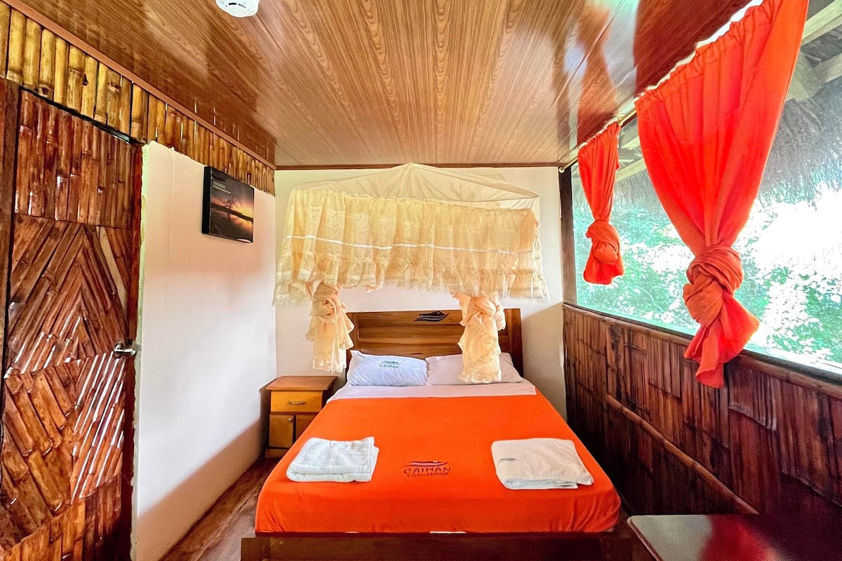 Een kamer met houten lambrisering, een bed, een nachtkastje en een raam met rode gordijnen.