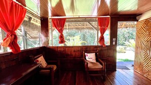 Un intérieur en bois avec un banc et des chaises, une fenêtre ornée de rideaux rouges et une vue sur la verdure à l’extérieur.