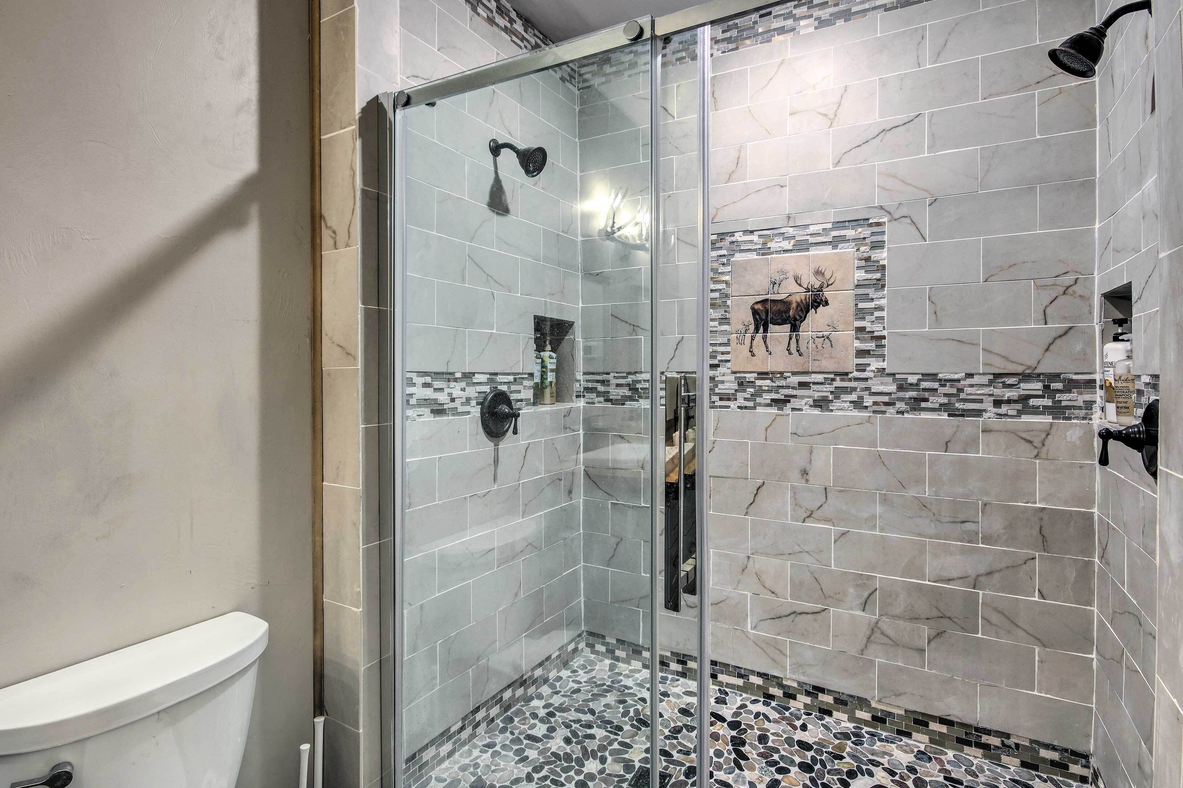 Une douche fermée par des parois de verre, avec un mur recouvert de carreaux de marbre, des toilettes et un sol en mosaïque.