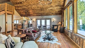 A spacious living room with a high ceiling, wooden beams, a chandelier, a leather sofa, a glass coffee table, and a large window offering a view of the outdoors. - Bozeman Cabin: Deck w/ Jacuzzi & Gas Grill! (Bozeman)
