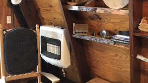 Una habitación cálida en el ático, con techo inclinado, un radiador antiguo, una silla, un taburete pequeño y estanterías llenas de libros y objetos decorativos.