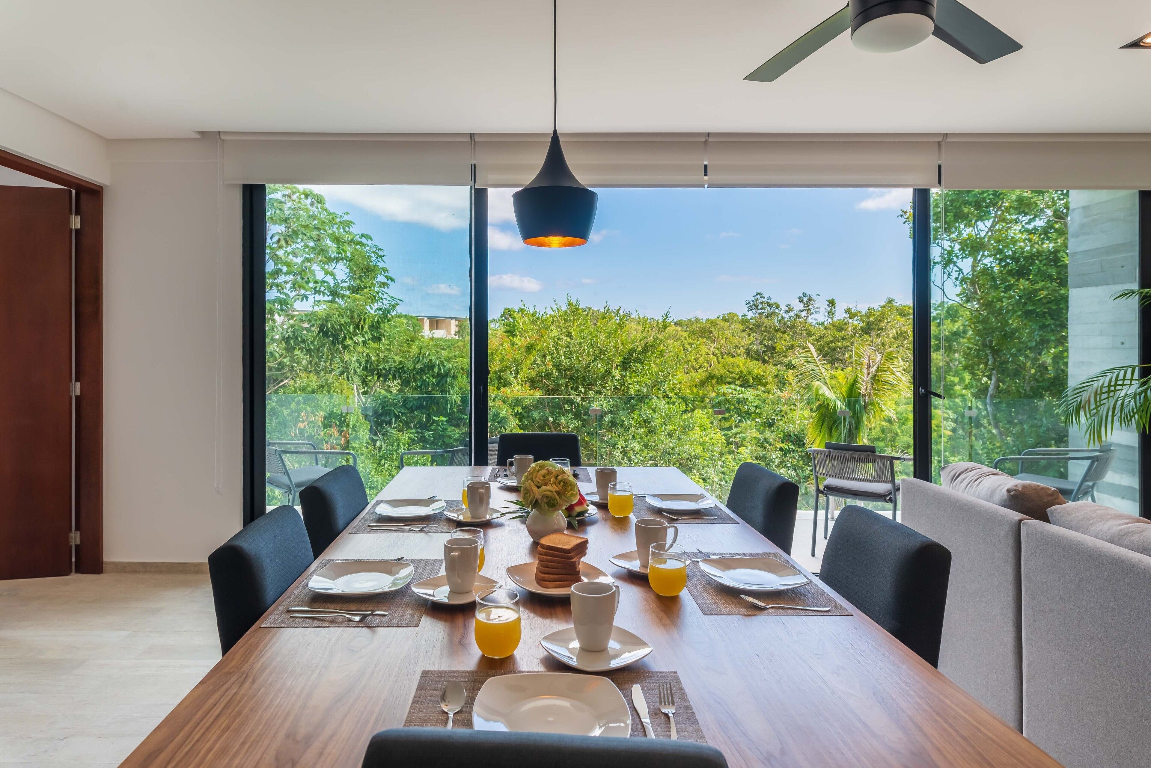 A dining area with a long table set for four, large windows, and a view of greenery.