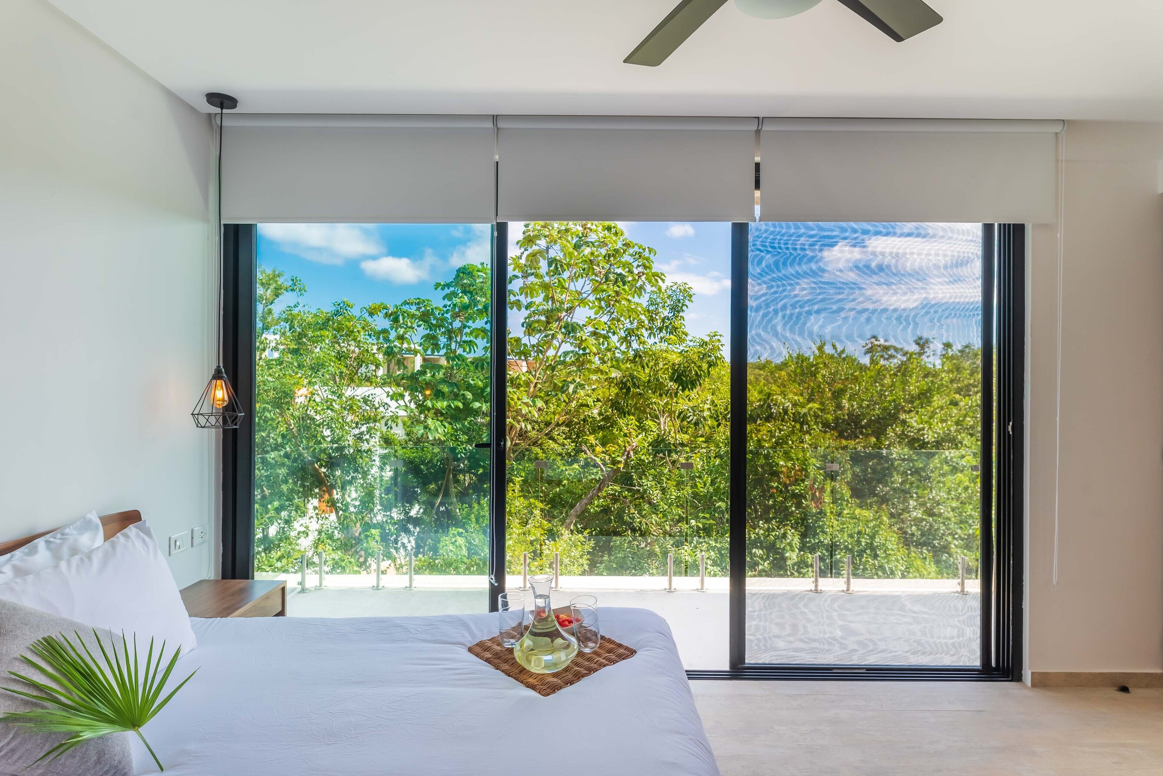 A modern bedroom with a large window, a bed with white linens, a bedside lamp, and a tray with a drink and fruit.