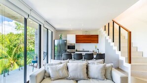A modern living room with a sofa, a kitchen area with a dining set, and a staircase. - Panoramic By Alegria Rentals and Sandy Powerful  (Tulum)
