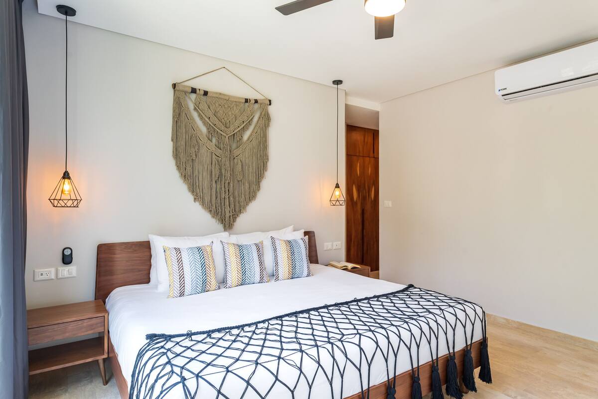 Quarto com cama, mesas de cabeceira, um pendente de macramé e um ventilador de teto.