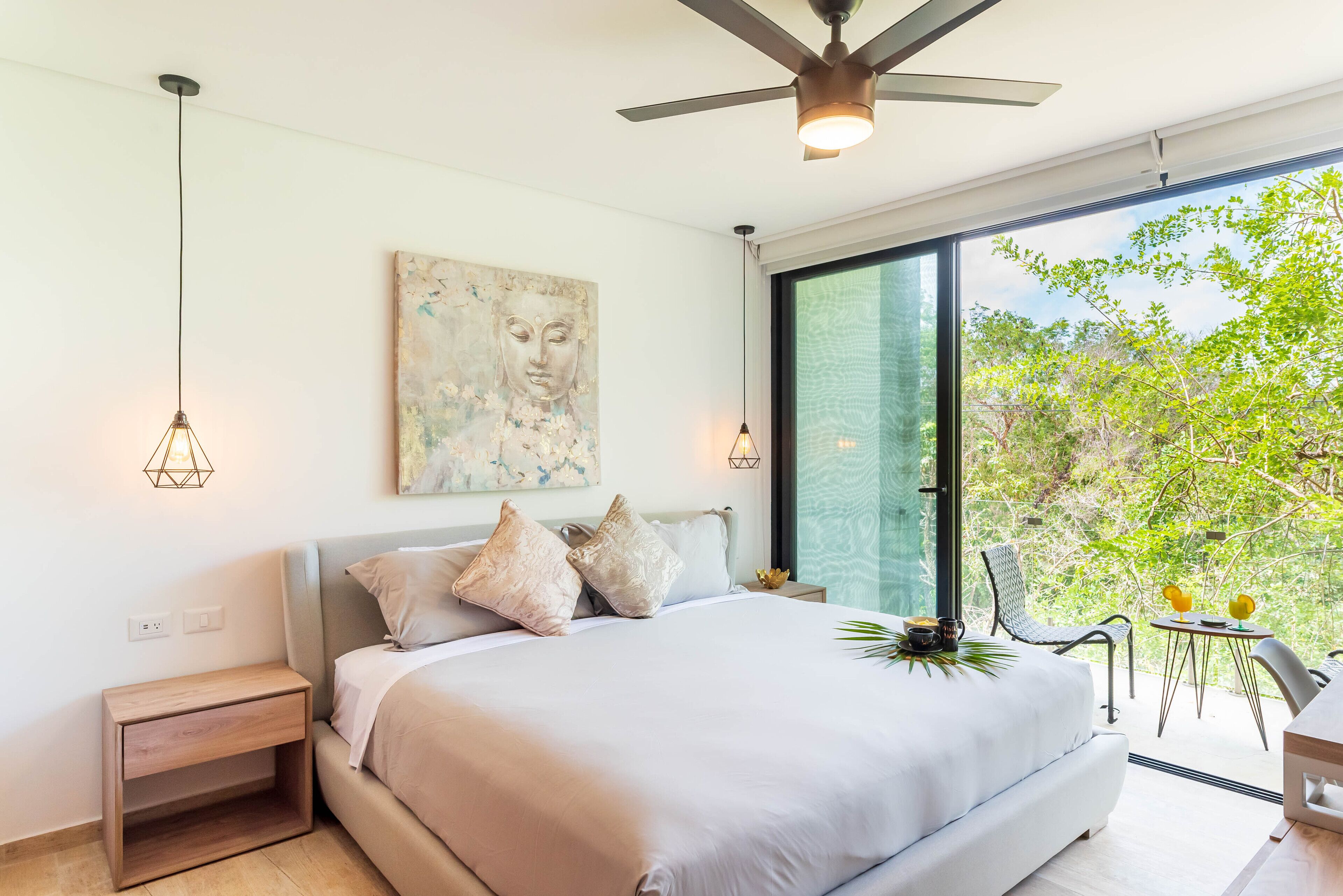 A modern bedroom with a large bed, a ceiling fan, and a view of greenery through a large window.