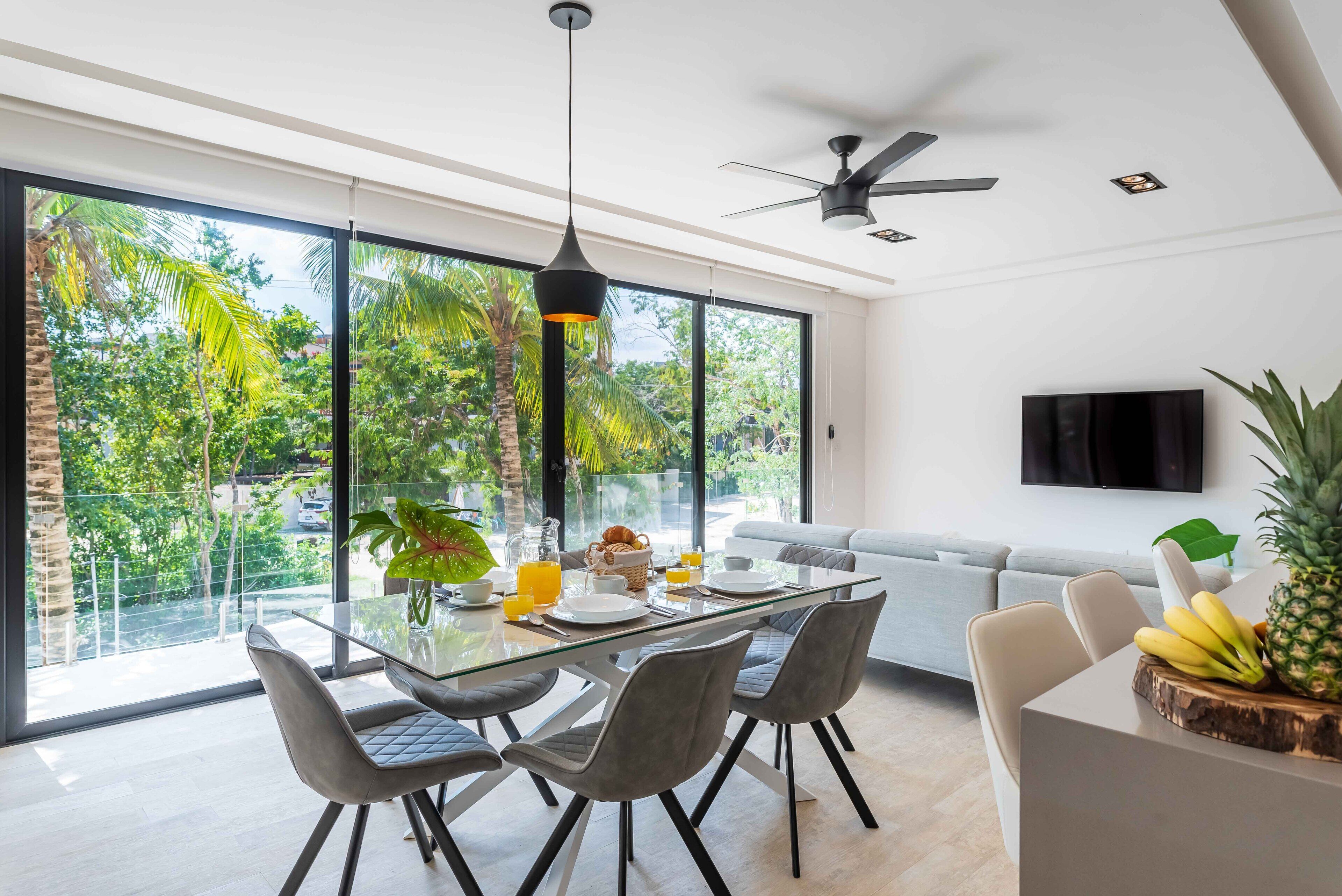 A modern dining area with a glass table, grey chairs, and a ceiling fan.