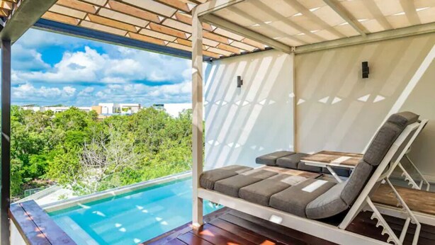 A poolside lounge area with a pergola overhead, a pool, and a view of greenery and buildings. - Panoramic By Alegria Rentals and Sandy Powerful  (Tulum)