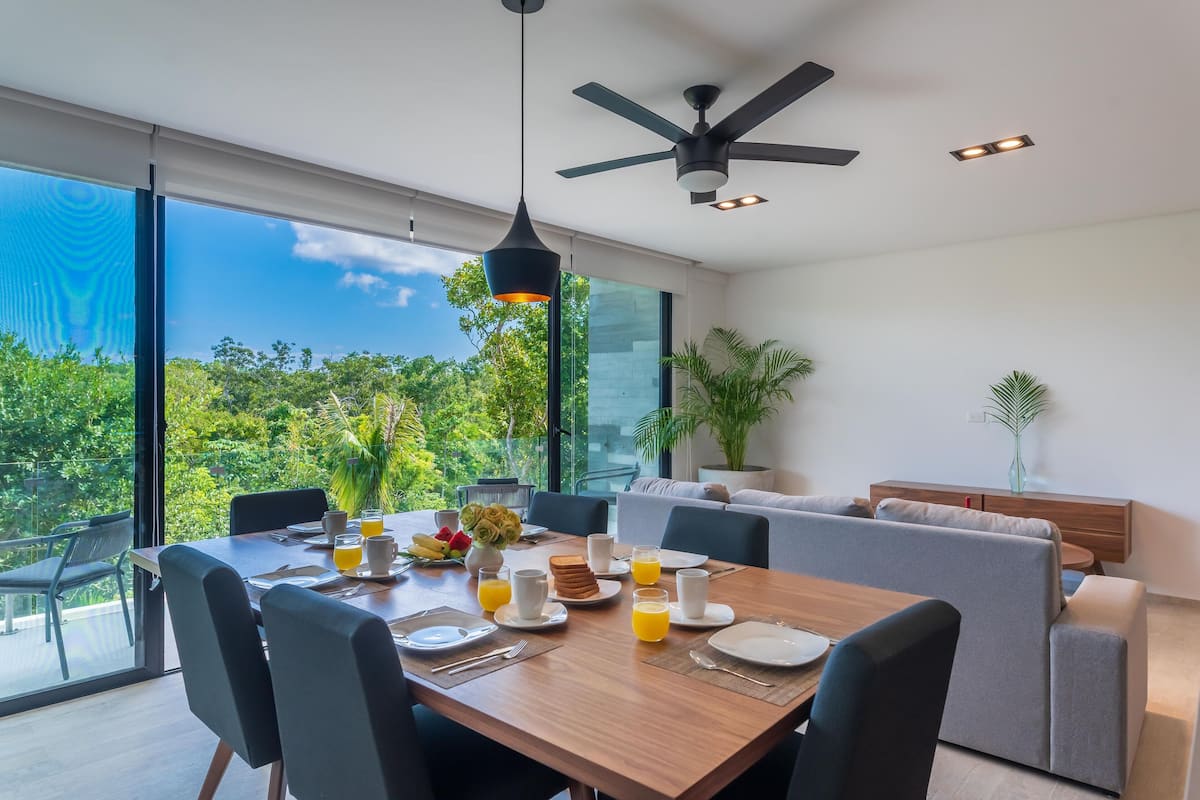 Uma sala de jantar moderna com uma mesa de madeira posta para o café da manhã, grandes janelas com vista para o verde e um ventilador de teto.