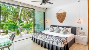 A modern bedroom with a large bed, a ceiling fan, a wall-mounted macrame piece, bedside lamps, and a view of a lush garden through a sliding glass door. - Panoramic By Alegria Rentals and Sandy Powerful  (Tulum)