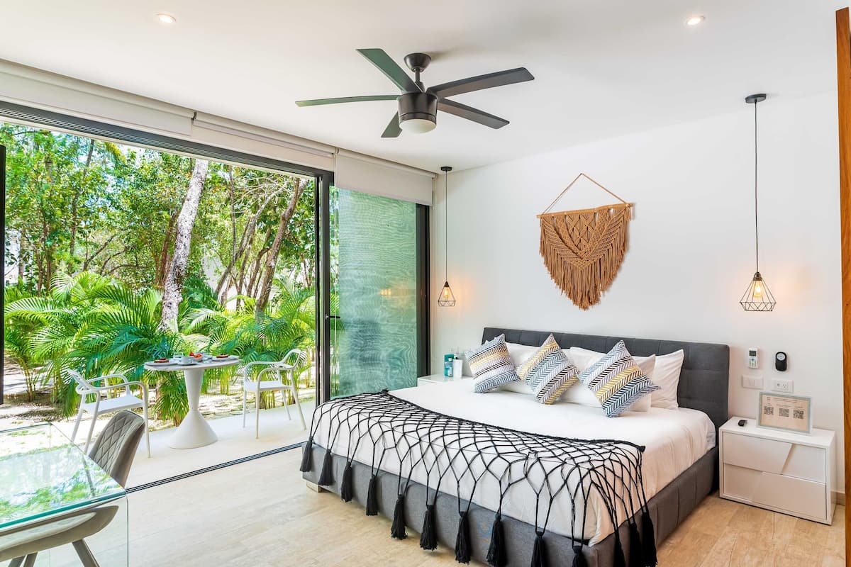 Quarto moderno com uma cama grande, um ventilador de teto, uma peça de macramé na parede, luminárias de cabeceira e vista para um jardim exuberante através de uma porta de vidro deslizante.