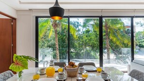 A dining table set for breakfast with a view of tropical trees through large windows. - Panoramic By Alegria Rentals and Sandy Powerful  (Tulum)