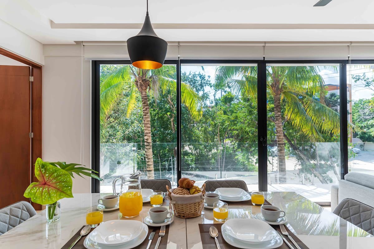 Mesa de jantar posta para o café da manhã, com vista para árvores tropicais através de grandes janelas.