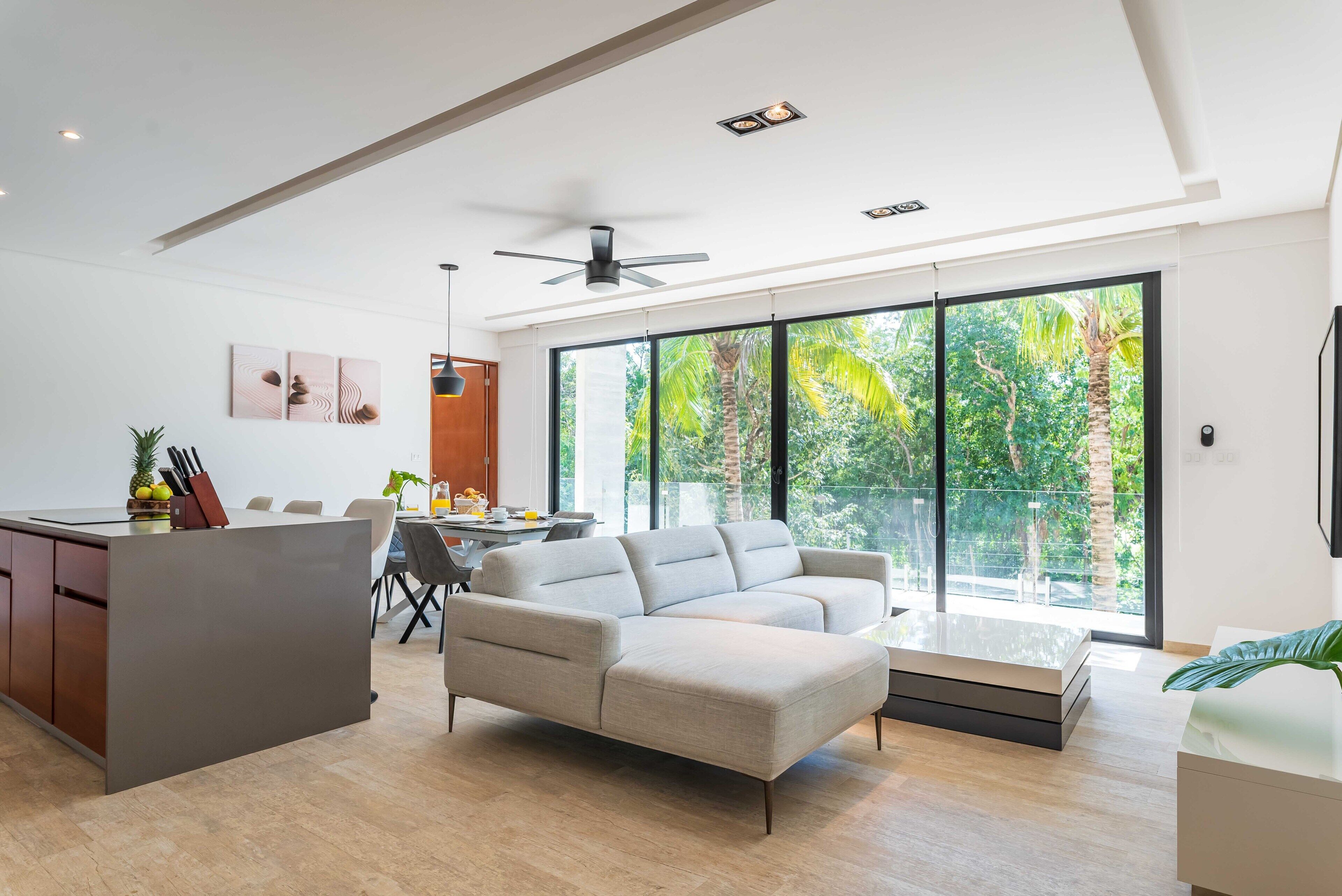 A modern living space with a sofa, dining area, and large windows offering a view of greenery.