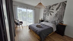 A bedroom with a bed, a chair, a nightstand, a wall art of palm trees, and a window with curtains.