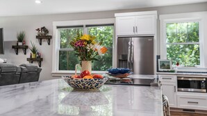 Une cuisine moderne avec des armoires blanches, un réfrigérateur en acier inoxydable, un îlot en marbre agrémenté d’un saladier de fruits et d’un vase de fleurs, et de grandes fenêtres offrant une vue sur la verdure.