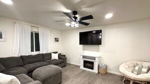 A living room with a sectional sofa, a television mounted on the wall, a ceiling fan, and a fireplace. - Prime location, Aurora viewing, Fast WI-FI!! (Fairbanks)