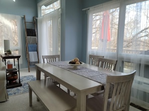 Uma sala de jantar com mesa de madeira, cadeiras e uma tigela de comida. Há uma escada e uma pequena mesa com uma planta e um livro.