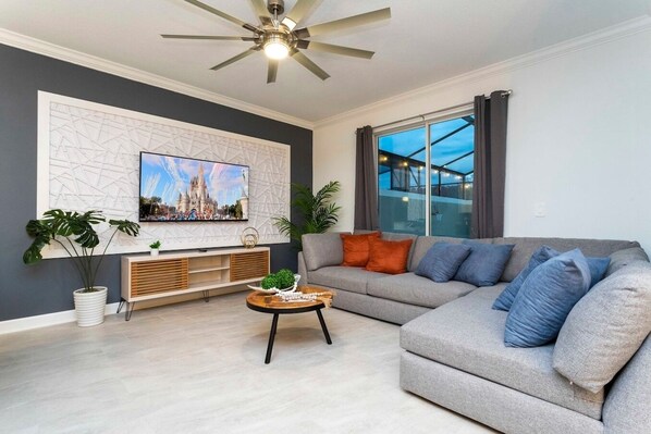A modern living room with a grey sofa, a coffee table, a TV mounted on a geometric wall, and a ceiling fan.