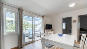 Une pièce moderne et bien éclairée, comprenant une table à manger, des chaises, un téléviseur et un balcon aménagé avec du mobilier d’extérieur.