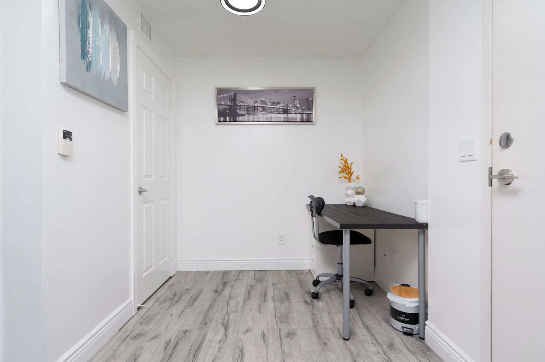 A clean, modern hallway with a wooden floor, a desk with a chair, a step-by-step trash bin, and a framed picture on the wall.