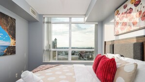A hotel room with a large window, a bed with white and red bedding, and a painting on the wall. - Duke Of York Boutique Suites (Mississauga)