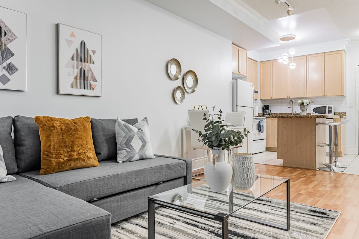 A modern living room with a grey sofa, a glass coffee table, and a kitchen in the background.