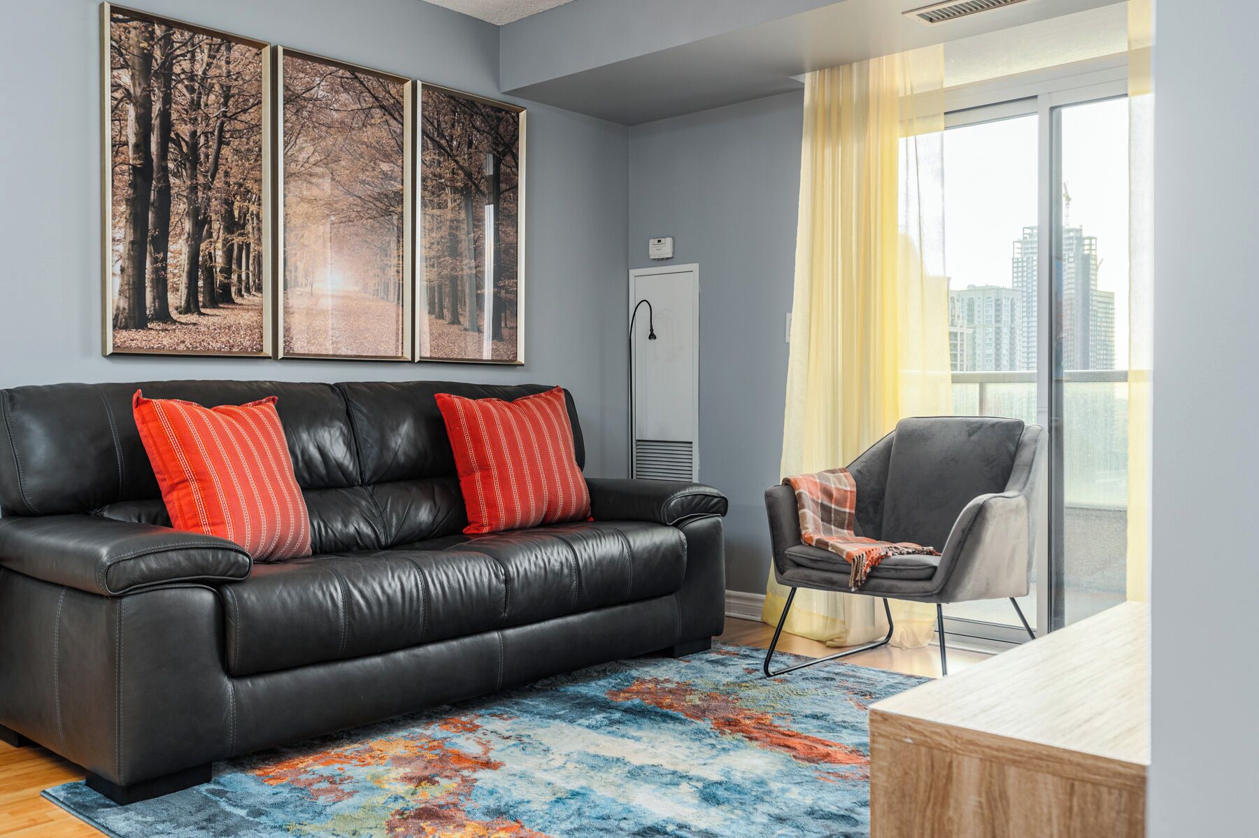 A modern living room with a black leather sofa, red pillows, a colorful rug, a grey armchair, and a large window with a city view.