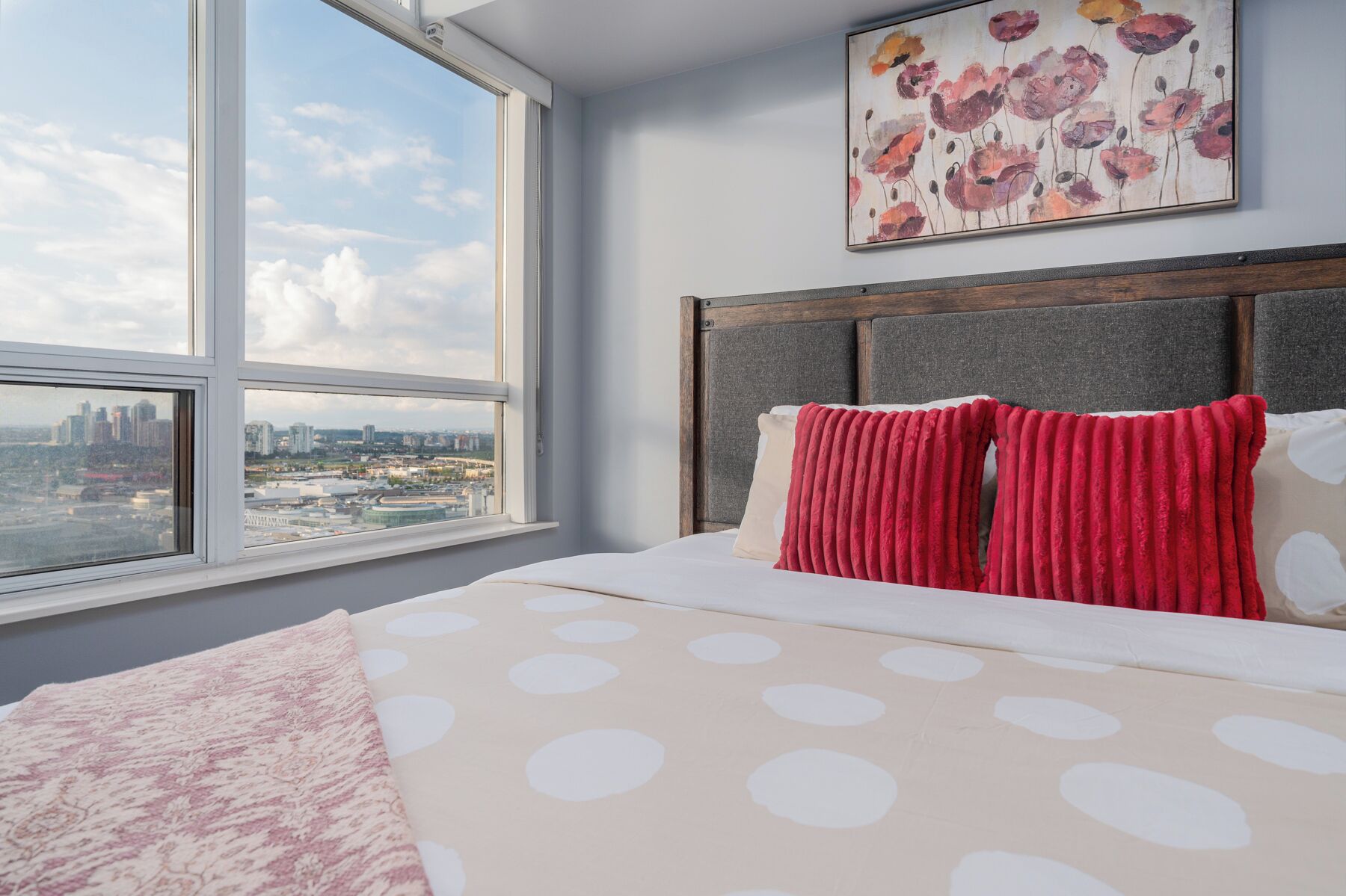 A bedroom with a large window, a bed with polka dot bedding, and a floral painting on the wall.