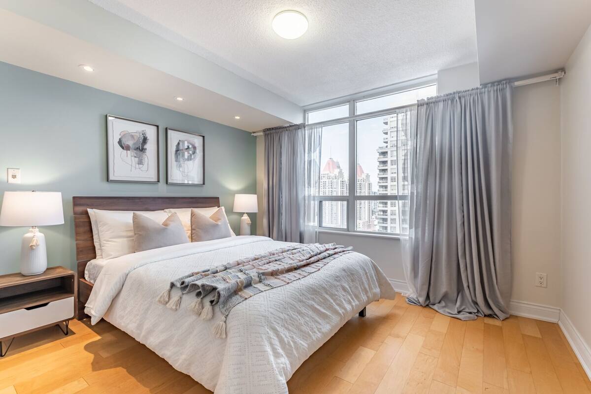 A modern hotel room with a large bed, two bedside tables with lamps, a window with curtains, and artwork on the wall.