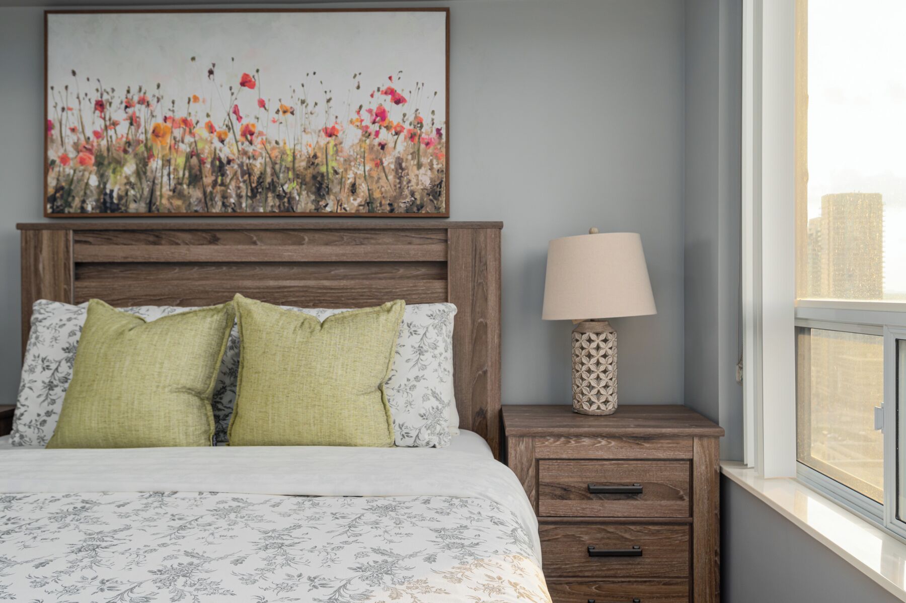 A bedroom with a wooden headboard, floral bedding, a bedside lamp, and a city view through a window.