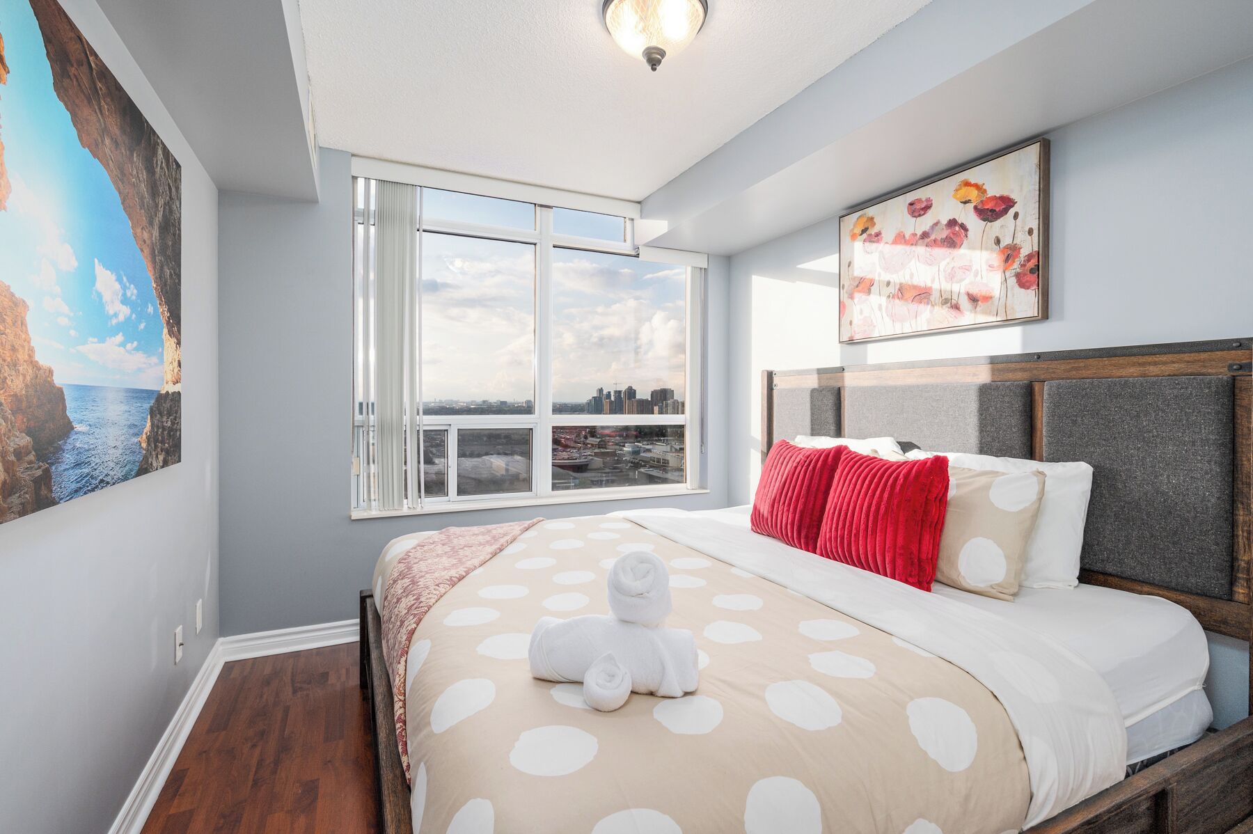 A bedroom with a bed, a window with a city view, and a painting on the wall.