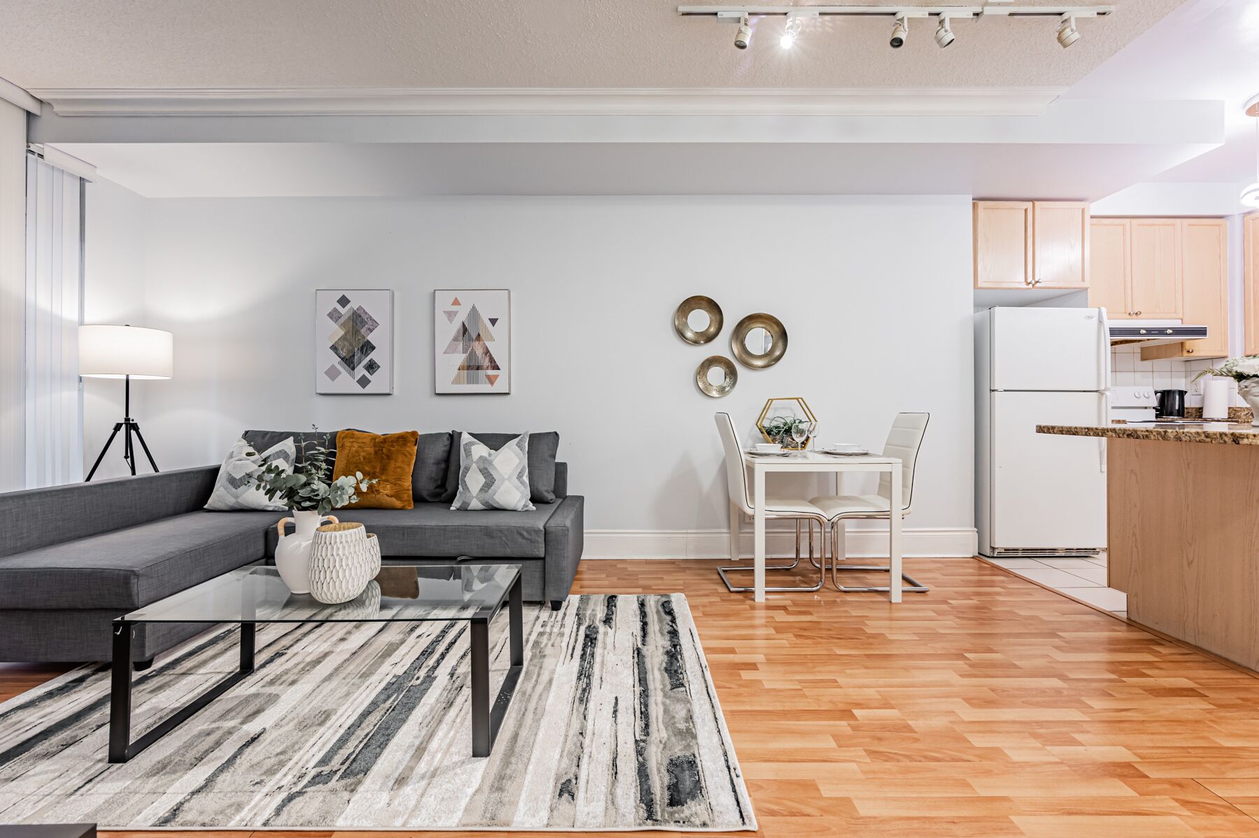 A modern living room with a grey sofa, a glass coffee table, and a dining area with a white table and chairs. A kitchen with wooden cabinets and a white refrigerator is visible in the background.