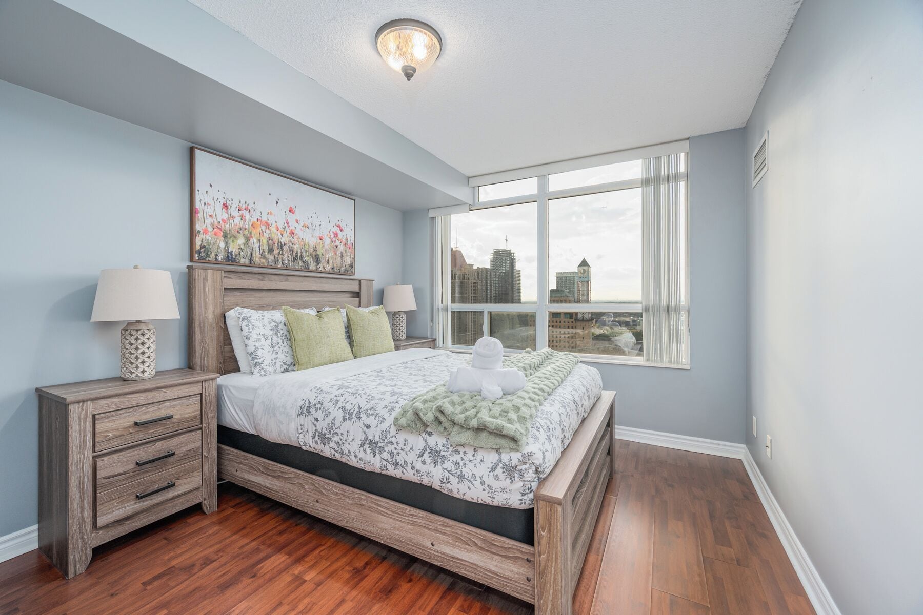 Un dormitorio con cama de madera, una mesita de noche con lámpara, vista a la ciudad a través de ventanas amplias y una lámpara de techo.