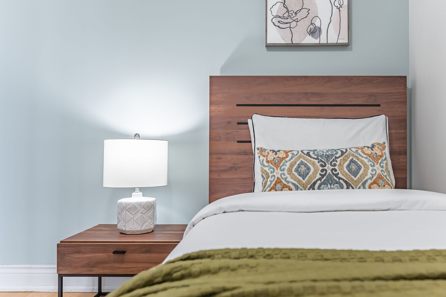 A neatly made bed with a patterned pillow, a bedside lamp, and a small white container on a wooden nightstand.