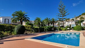 Una piscina con borde de piedra, rodeada de palmeras y una ladera con edificios blancos con balcones.