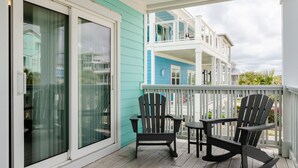 A covered porch with two Adirondack chairs and a small table, overlooking a residential area with a blue house in the background. - Spacious 5 BR Home Across from Beach! (Kure Beach)