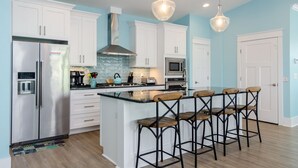 A modern kitchen with white cabinets, a stainless steel refrigerator, a built-in oven, and a central island with bar stools. - Spacious 5 BR Home Across from Beach! (Kure Beach)