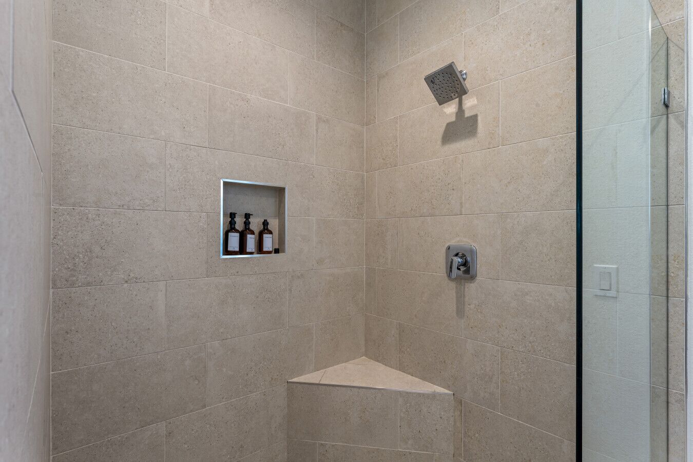 A shower area with beige tiles, a glass enclosure, and a shelf with three bottles.