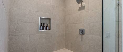 A shower area with beige tiles, a glass enclosure, and a shelf with three bottles.