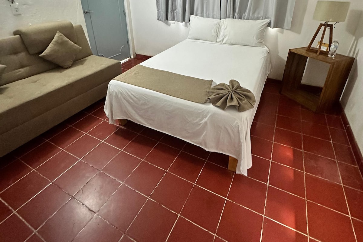 A hotel room with a bed, a sofa, a wooden nightstand, a lamp, a ceiling fan, and a door.