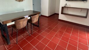 Una habitación con piso de baldosas rojas, una mesa de madera con sillas, una puerta azul y un televisor montado en la pared.